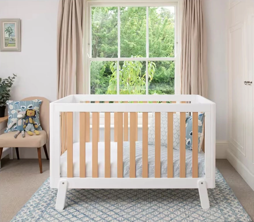 White wooden cot bed with natural timber slats and angled feet in a bright nursery, with a window and a blue patterned rug White wooden cot bed with natural timber slats and angled feet in a bright nursery, with a window and a blue patterned rug